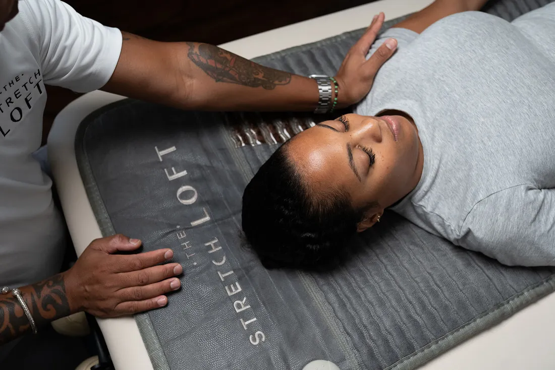 Overhead view of a client lying on The Stretch Loft mat, eyes closed, as her practitioner cradles her head