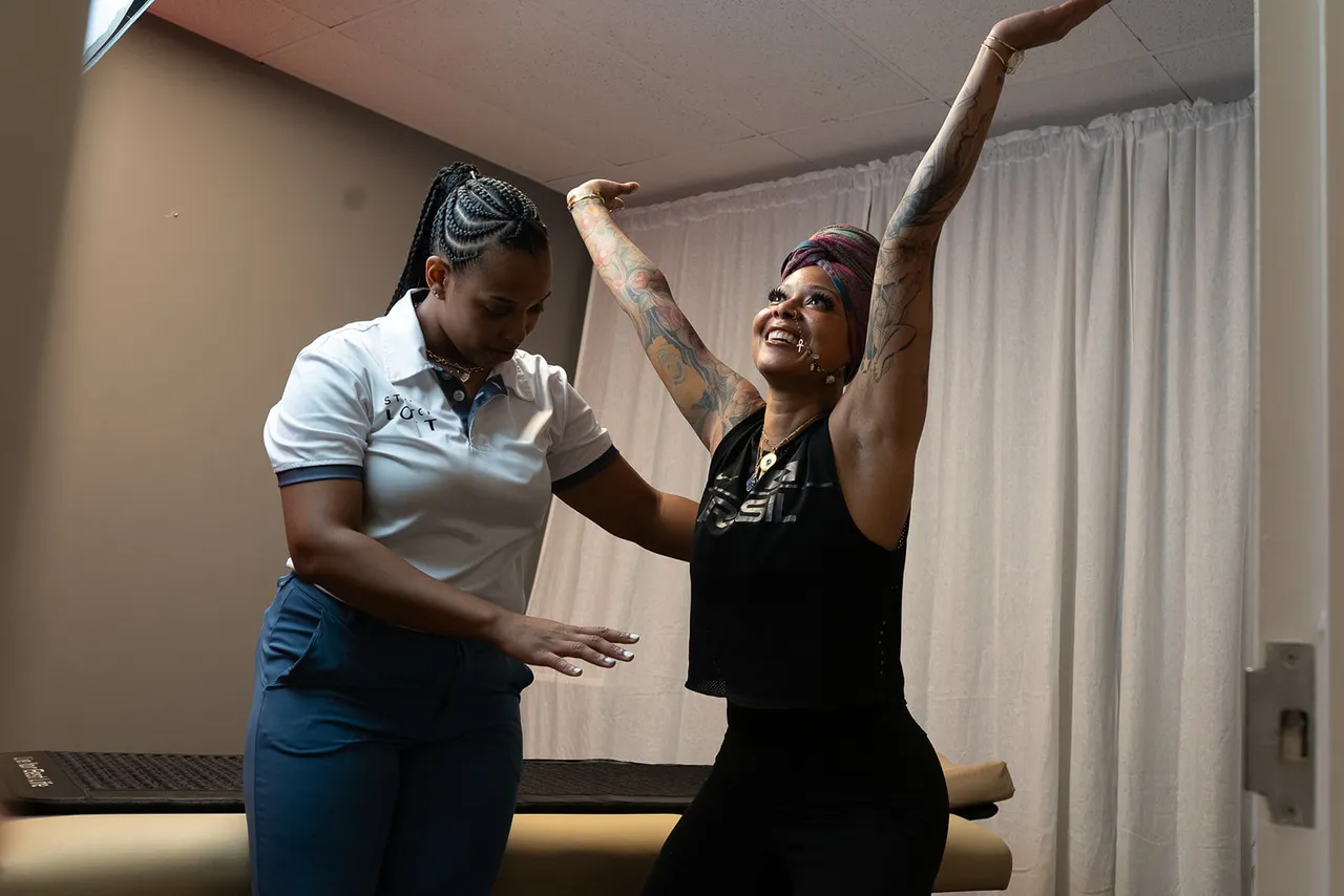 Client reaching both arms overhead and smiling as a practitioner stands beside her in the studio